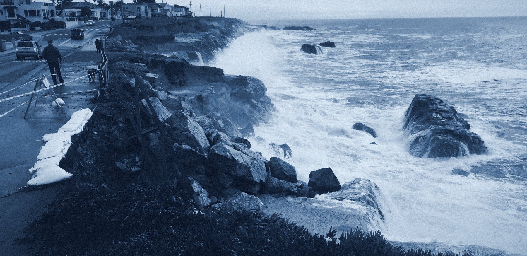 Storm waves breaking over the riprap at the foot of an eroding bluff beside East Cliff Drive in Santa Cruz, California, in January 2016, with the roadside taped off, sandbags along the edge and people watching.